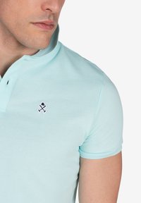 Camisa polo azul claro, hecha de tela texturizada. Presenta una tapeta de dos botones y un pequeño logotipo bordado en el lado izquierdo del pecho.