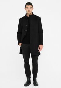 Black wool coat with a high collar and six buttons, worn over a textured black sweater and fitted black trousers, paired with black ankle boots.