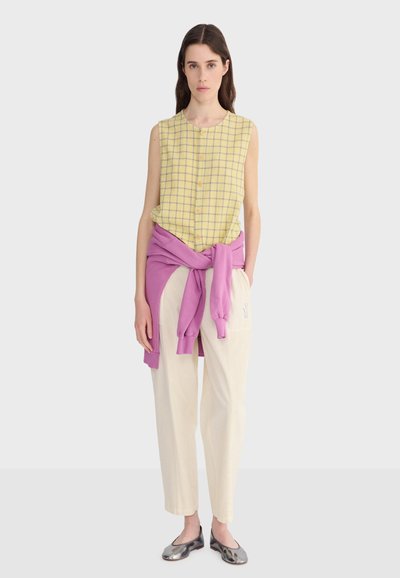 Jeune femme portant un chemisier sans manches à carreaux jaunes, un pantalon crème, des ballerines métalliques, et un sweat-shirt rose noué autour de la taille, debout devant un fond uni.