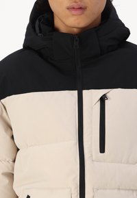 Puffermantel mit der oberen Hälfte in Schwarz und der unteren Hälfte in Beige, mit einem Frontreißverschluss, verstellbarer Kapuze und einer Brusttasche mit Reißverschluss.