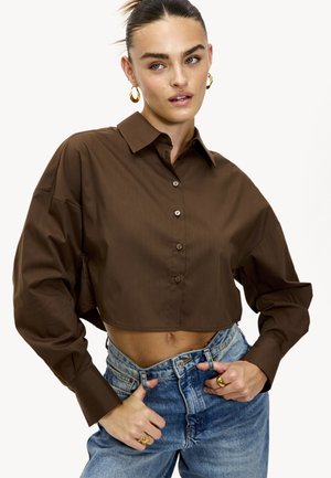 Chemise marron courte en tissu lisse, dotée d'un devant boutonné, d'un col et de manches longues. Associée à un jean taille haute bleu clair.