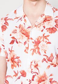 Camicia a maniche corte in tessuto bianco con un motivo floreale colorato in tonalità di rosa, arancione e rosso. Colletto classico e chiusura frontale con bottoni.