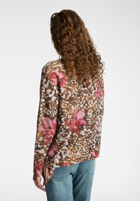 Donna con lunghi capelli ricci che indossa un maglione marrone a fantasia floreale e jeans blu, vista di spalle su uno sfondo neutro.