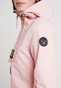 Jasnoróżowa kurtka z kapturem, zapinana na suwak przy szyi, z przednią kieszenią i okrągłą czarną naszywką na rękawie. Gładka faktura, casualowy design.