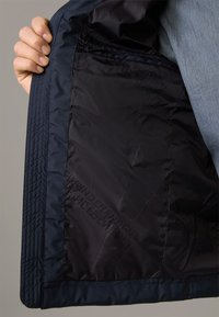 Main tenant ouverte la doublure intérieure sombre d'une veste marine, montrant une poche zippée et un texte subtilement imprimé sur le tissu.