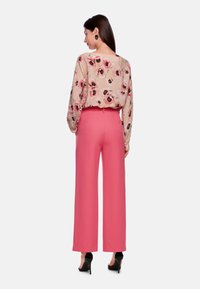 Blumenbluse in Beige mit rosa und schwarzen Akzenten, kombiniert mit hochtaillierten, weit geschnittenen rosa Hosen und schwarzen High-Heels.