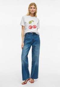T-shirt blanc avec un graphisme de cerise, associé à un jean bleu taille haute à jambes larges et des sandales à talons rouges. Fond uni.