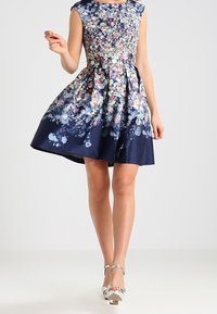 Blumenkleid mit rundem Ausschnitt, ärmellos, marineblauem Grund, verschiedenen bunten Blumenmustern und einem ausgestellten Rock. Sichtbare silberne Knöchelriemen.