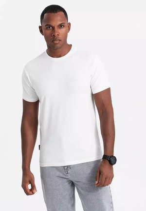 White cotton T-shirt featuring a crew neck and short sleeves. Simple design with a small logo detail on the chest. Worn with grey jeans.