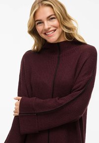 Maglione lavorato a maglia borgogna con colletto alto, vestibilità morbida, texture a coste e dettagli neri lungo le cuciture laterali e sui polsini.