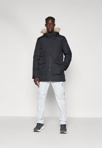 Μαύρο μπουφάν με κουκούλα επενδεδυμένη με τεχνητή γούνα, που διαθέτει δύο μπροστινές τσέπες και κλείσιμο με φερμουάρ. Συνδυασμένο με ανοιχτό γκρι cargo παντελόνι και μαύρα sneakers.