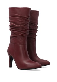 Pár burgundských kožených bot po lýtka se volným holenním dílem, špičatými špičkami a štíhlými vysokými podpatky na bílém pozadí.