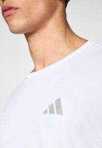 Biała sportowa koszulka z krótkim rękawem wykonana z teksturowanej tkaniny, z srebrnym logo z trzema paskami na lewym górnym ramieniu.