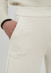 Pantalone boje kreme s uzorkom houndstooth, s glatkom teksturom i bočnim džepovima. Pojas je strukturiran i ravan.