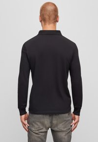 Svart långärmad polo med klassisk krage och ribbade ärmar, tillverkad av ett slätt tyg, visas från baksidan. Grå jeans syns nedanför.