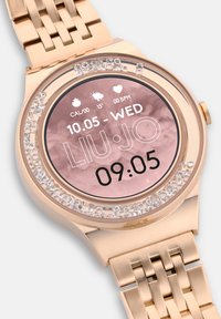 Smartwatch color oro con quadrante rotondo, display marmorizzato rosa, accenti in cristallo attorno al bezello e bracciale metallico a maglie.