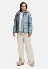 Giacca a vento azzurro chiaro con cappuccio, chiusura con zip e dettagli di cucitura orizzontale. Abbinata a pantaloni di velluto a coste beige e scarpe beige.