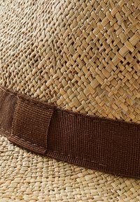 Olkihattu, jossa on kudottu pinta luonnollisissa sävyissä, ja siinä on ruskea kangasnauha pohjan ympärillä. Muoto on pyöreä ja leveä lierikanta.