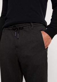 Housut harmaalla herringbone-kuviolla, jossa on naru vyötäröllä ja sivutaskut, valmistettu teksturoidusta kankaasta. Yhdistetty mustaan villapaitaan.