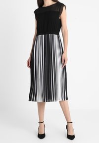 Schwarz-weiß gestreiftes Kleid mit einem transparenten schwarzen Oberteil, kurzen Ärmeln und einem plissierten Rock. Akzentuiert mit schwarzen Absätzen.