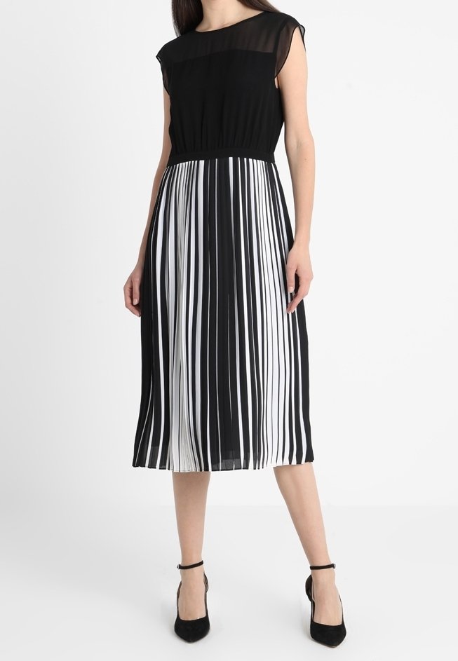 Schwarz-weiß gestreiftes Kleid mit einem transparenten schwarzen Oberteil, kurzen Ärmeln und einem plissierten Rock. Akzentuiert mit schwarzen Absätzen.