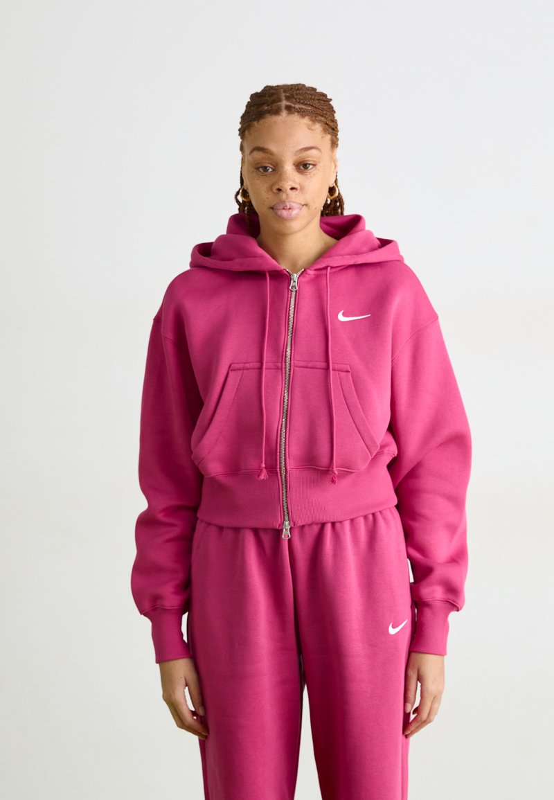 Ružová mikina na zips vyrobená z mäkkej tkaniny, so kengurským vreckom, šnúrkami a malým bielym logom Nike na hrudi. V páre s odpovedajúcimi teplákmi.