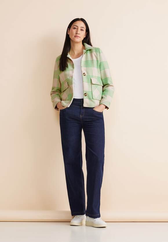 Giacca a quadri verde e crema con due tasche frontali, abbinata a jeans in denim scuro e sneakers bianche, ambientata su uno sfondo neutro.