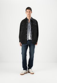 Chaqueta de pana negra sobre una camisa a cuadros azul y blanca, combinada con jeans oscuros y zapatillas blancas. Diseño simple y corte clásico.