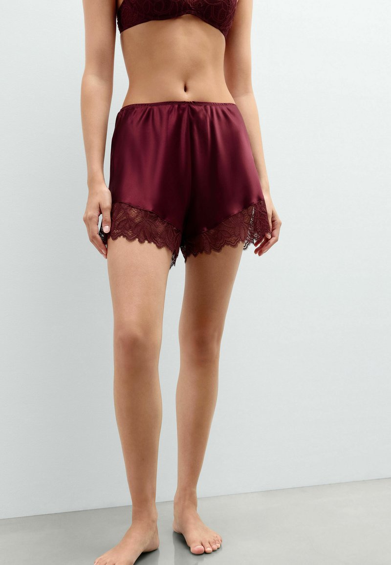 Woman wearing burgundy satin shorts with lace trim and matching bra, standing barefoot against a plain light gray background.