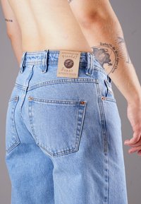 Light blue denim jeans with a high waistband, prominent back pockets, and a tan leather tag labeled "Picalde Jeans Authentic Design."