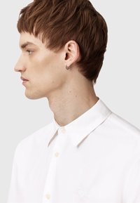 Camisa blanca de botonadura completa con cuello de punta y pequeño logo de corazón bordado en el pecho. Lleva puesta una persona con cabello corto y castaño.