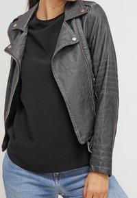 Giacca da moto nera in ecopelle con colletto strutturato, accessori in argento e dettagli a coste sulle maniche. Indossata sopra un top nero.