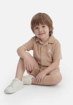 Νεαρό αγόρι με ανοιχτό καστανά μαλλιά που κάθεται με σταυροπόδι, φοράει μια μπεζ πουκαμίσα με γιακά και κοντά παντελόνια με λευκά αθλητικά παπούτσια.