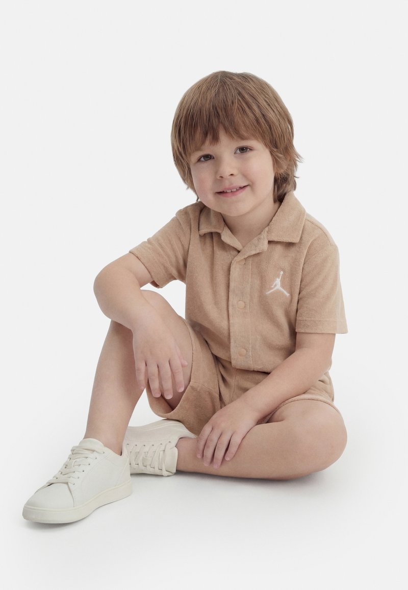 Νεαρό αγόρι με ανοιχτό καστανά μαλλιά που κάθεται με σταυροπόδι, φοράει μια μπεζ πουκαμίσα με γιακά και κοντά παντελόνια με λευκά αθλητικά παπούτσια.