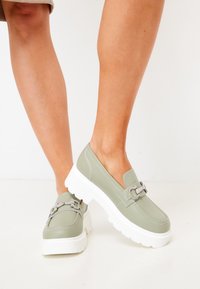 Gröna faux läder slip-on skor med en tjock vit sula, prydda med en dekorativ silverkedja på ovandelen och subtila sömdetaljer.