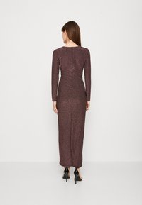 Robe longue ajustée dans un bordeaux foncé avec un tissu texturé et scintillant. Dotée de longues manches et d'une fermeture éclair centrale au dos. Des talons noirs sont portés.