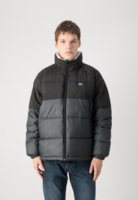Giacca a vento imbottita con parte superiore nera e sezione inferiore grigia, colletto alto, chiusura con zip e polsini elastici, realizzata in materiale lucido e isolante.