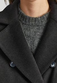 Abrigo peacoat oscuro de lana con solapas con muescas, doble botonadura y un suéter de punto gris suave debajo, mostrando texturas y tonos ricos.