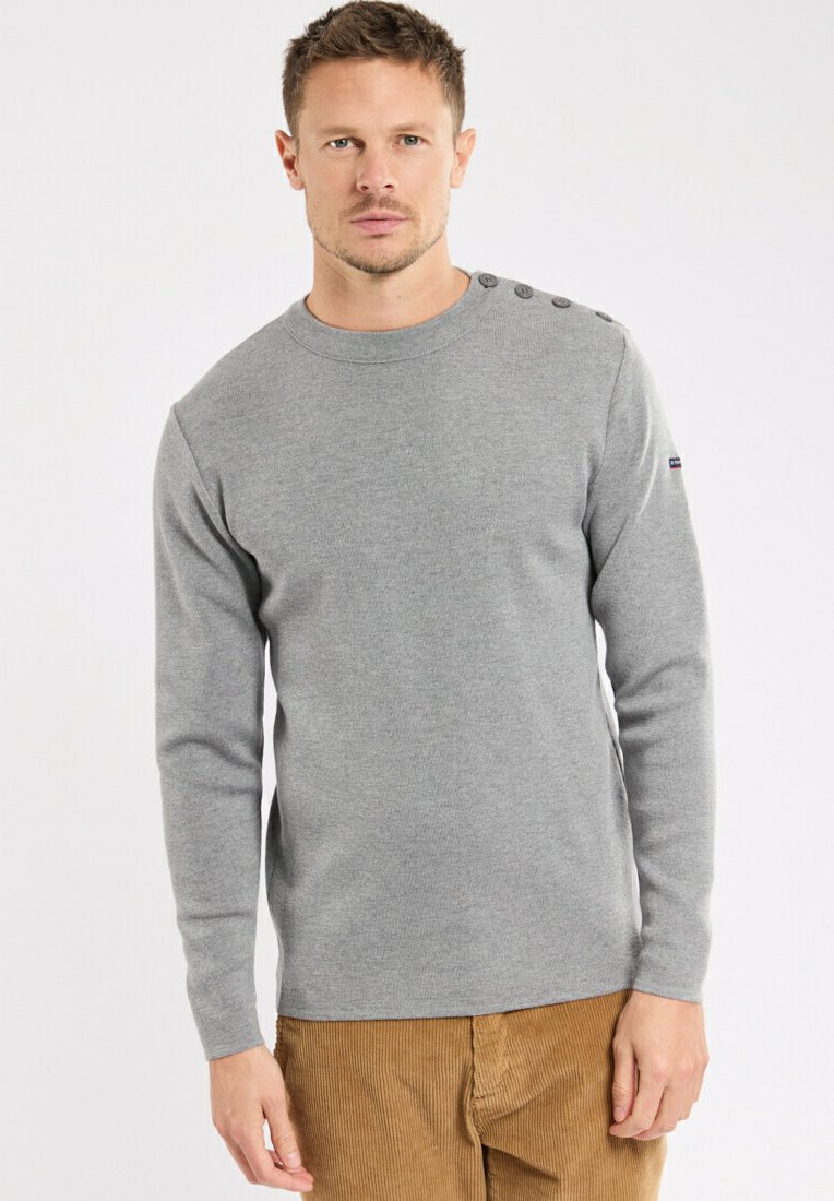 Maglione grigio a maniche lunghe con scollo rotondo e dettaglio a quattro bottoni su una spalla, realizzato in un tessuto morbido. Il modello indossa pantaloni in velluto.