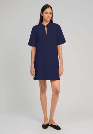 Femme debout devant un fond gris portant une robe bleu marine à manches courtes avec col et des chaussures noires sans lacets.