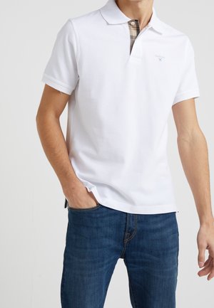Biała koszulka polo z krótkim rękawem, klasyczny kołnierzyk oraz kratkowy detal wewnątrz kołnierza. Nosi się z ciemnoniebieskimi dżinsami, o gładkiej bawełnianej strukturze.