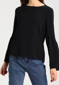 Top nero a maniche lunghe con scollatura rotonda e maniche a campana, realizzato in tessuto leggero. Abbinato a jeans blu.