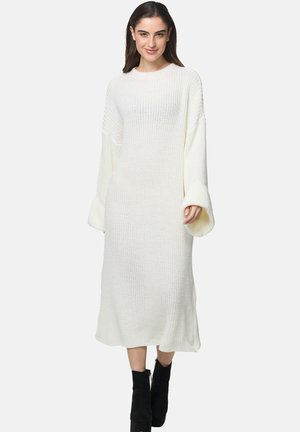 Vestido blanco de punto de manga larga con corte holgado, patrón texturizado y puños anchos, combinado con botas negras hasta el tobillo.