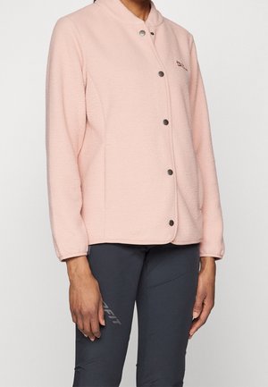 Nainen pukeutuneena vaaleanpunaiseen fleece-takkiin, jossa painonapit, yhdistetty tummanharmaisiin istuviin housuihin yksiväristä taustaa vasten.