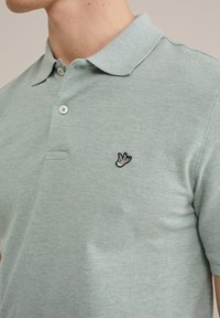 Svetlozelena polo majica z rahlo teksturo, s tri gumbi na zapenjanju in črnim vezenim logotipom ptice na prsi.