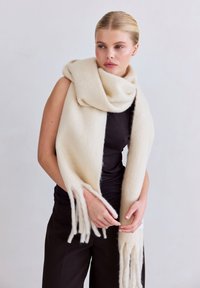 Beige wollen sjaal met franjes aan de uiteinden, losjes om de schouders gedrapeerd. Zachte structuur, grote rechthoekige vorm. Glad, minimalistisch ontwerp.