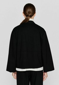 Zwarte oversized jas met brede mouwen en een relaxte pasvorm, voorzien van een naaddetail dat horizontaal over de achterkant loopt. Gladde textuur.