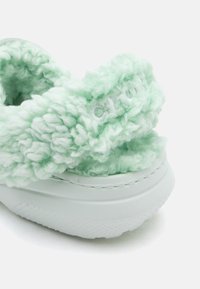 Chaussure en vert clair duveteux avec une texture en peluche et une semelle en caoutchouc blanche, gros plan sur la zone du talon montrant le matériau doux et moelleux ainsi que les détails de couture.