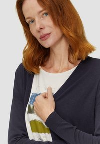 Cardigan bleu marine à manches longues, décolleté en V et poignets côtelés, porté sur un t-shirt léger avec un imprimé graphique multicolore.