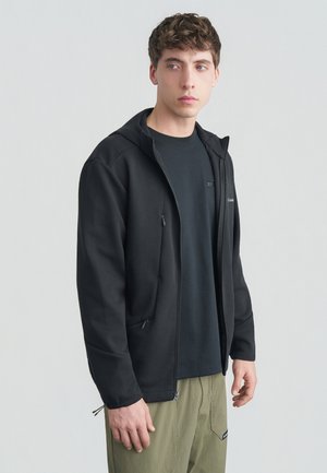 Giovane uomo che indossa una felpa nera con cerniera sopra una maglia scura e pantaloni verde oliva, in piedi contro uno sfondo grigio chiaro uniforme, guarda a sinistra.